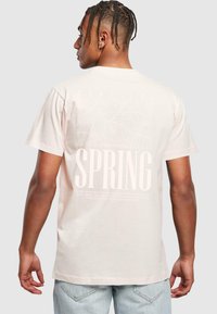 Vaaleanpunainen puuvillateeppari, jonka selässä on suuri valkoinen "SPRING" -printti ja trooppinen lehtigrafiikka, lyhyet hihat ja pyöreä kaula-aukko.