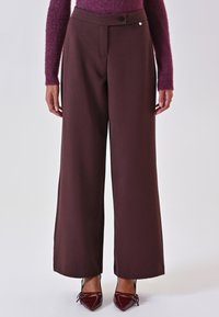 Pantaloni a gamba larga in un profondo tessuto bordeaux, con chiusura a bottone e un sottile accento metallico. Texture liscia, design su misura.