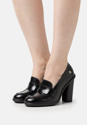 Mocassins à talons hauts en cuir verni noir avec une sangle décorative sur le dessus et un talon carré texturé. Agrémentés d'un petit détail de logo.