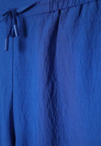 Pantalon en tissu bleu avec une ceinture élastique et un cordon de serrage avec extrémités en plastique, montrant les détails de la texture et des coutures.