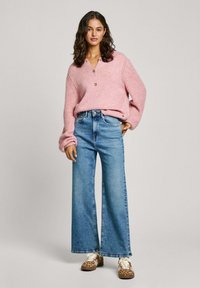 Suéter de punto rosa con escote en V y detalle de botones, combinado con jeans de pierna ancha azules y zapatos marrones con estampado.