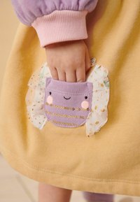 La main d'un enfant à l'intérieur d'une petite poche en tissu violet avec un visage souriant et des volants décoratifs sur un vêtement jaune.