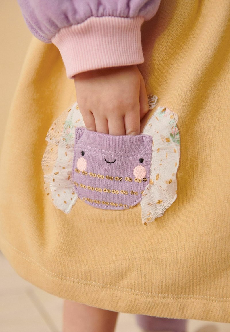 La main d'un enfant à l'intérieur d'une petite poche en tissu violet avec un visage souriant et des volants décoratifs sur un vêtement jaune.