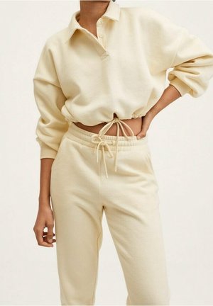 Personne portant un sweat-shirt court à manches longues crème avec col et un pantalon de survêtement assorti à cordon, une main sur la hanche et l'autre détendue.