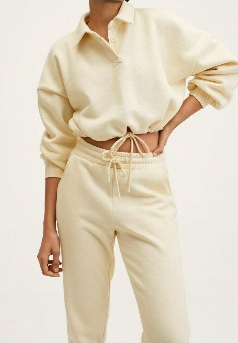 Personne portant un sweat-shirt court à manches longues crème avec col et un pantalon de survêtement assorti à cordon, une main sur la hanche et l'autre détendue.
