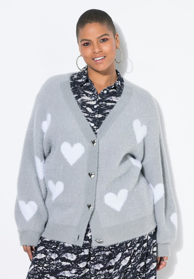 Persona sorridente con capelli corti indossa un cardigan grigio con cuori bianchi e bottoni a forma di cuore lucidi sopra una camicia a righe bianche e nere.
