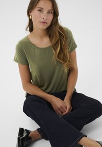 T-shirt verde oliva a maniche corte con scollatura rotonda abbinato a jeans larghi blu scuro. Il modello è seduto con le mani appoggiate sulle ginocchia.