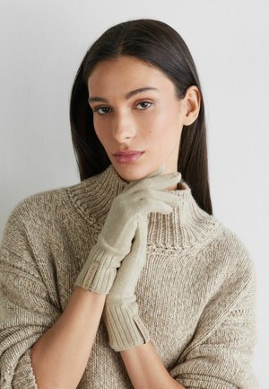 Beige stickade handskar med ribbade muddar och logotaggar, båda på händer och i kombination med en strukturerad beige tröja. Mjukt, varmt material.