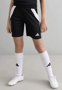 Melni sporta šorti ar baltām sānu svītrām, elastīga josta un Adidas logotips. Apvienoti ar baltām ceļgalu zeķēm un baltām sporta kurpēm.