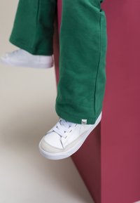 Witte sneaker met gestructureerde panelen en veters, gecombineerd met groene joggingsbroek. De broek heeft een label aan de zoom; beide kledingstukken liggen tegen een roze oppervlak.