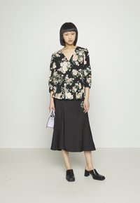 Blusa envolvente floral preta com flores em rosa e verde, combinada com uma saia A-line preta. O modelo segura uma clutch lavanda e está a usar mules pretas de salto bloco.