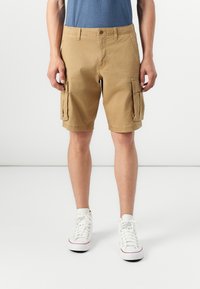Khaki szorty cargo o luźnym kroju, z bocznymi kieszeniami, zapięciem na guzik i paskami, wykonane z materiału bawełnianego. Połączone z białymi trampkami.