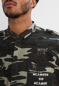 Camicia mimetica in verde scuro e nero, con collo a risvolto, tasche con patta e scritta bianca "SEASONS NO SEASON" con dettagli consumati.