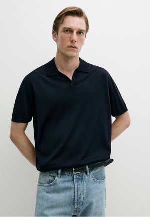 Jeune homme aux cheveux châtain clair portant une chemise noire à manches courtes avec col et un jean bleu clair sur un fond clair uni.