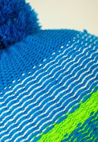 Knitted beanie in blue with white wavy stripes and a green accent. Features a fluffy blue pom-pom on top and textured fabric.