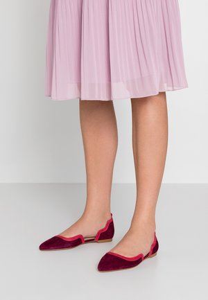 Bordeauxrote Wildleder-Ballerinas mit spitzer Kappe und geschwungenen Kanten, akzentuiert durch einen kontrastierenden pinkfarbenen Rand. Platziert auf einem hellen Hintergrund.