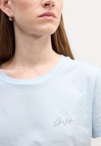 Svetlomodré bavlnené tričko s okrúhlym výstrihom, na ľavej strane s dizajnom z kryštálikov. Hladká textúra, bežný strih.