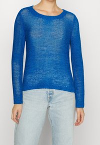 Kvinna som bär en genomskinlig, långärmad, kunglila stickad tröja kombinerad med ljus tvättade högmidjade jeans, stående mot en enkel bakgrund.