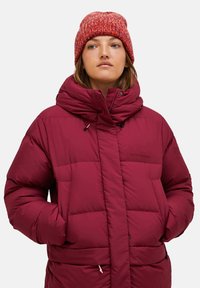 Vrouw met een rode gebreide beanie en een dikke bordeauxkleurige winterjas, met haar handen in de zakken, die iets opzij kijkt.