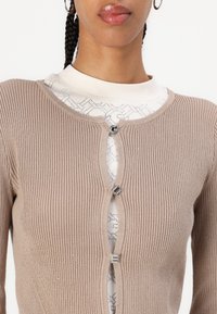Beige ribbed cardigan with three decorative silver buttons, layered over a white top featuring a subtle geometric pattern.