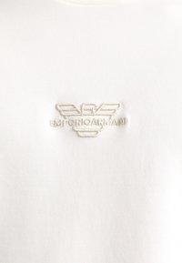 Camiseta blanca de algodón con un logo bordado de Emporio Armani en beige claro, con una textura suave y un cuello redondo clásico.