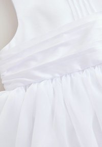 Robe blanche avec un corsage ajusté, texture plissée et une jupe en tulle évasée. Le matériau semble léger et lisse, mettant en valeur un design épuré.