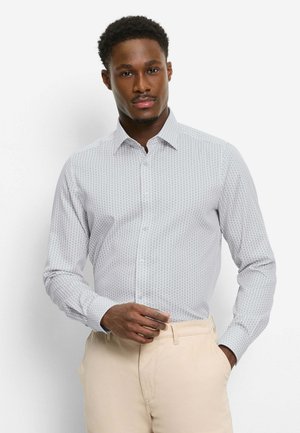 Mann trägt ein hellgraues, gemustertes Langarmhemd, in beigen Hosen gesteckt, steht mit einer Hand, die das Hemd richtet, und der anderen in der Tasche.