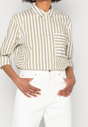 Person iført en beige og hvid vertikalt striped knap-op skjorte med en striped brystlomme og hvide højtaljede bukser med hænderne i lommerne.