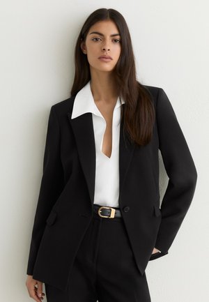 Mujer con cabello largo y oscuro que lleva un blazer negro, blusa blanca con escote en V profundo, pantalones negros y un cinturón negro con hebilla dorada.