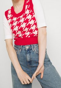 Dzianinowy top bez rękawów w wzór pepitki w kolorach czerwonym i kremowym, zestawiony z wysokim stanem niebieskich dżinsów z przecieranymi detalami.