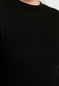 Schwarzer, gerippter Pullover mit rundem Halsausschnitt. Die Stoffstruktur zeigt vertikale Linien, und die Nahaufnahme konzentriert sich auf den Brustbereich.