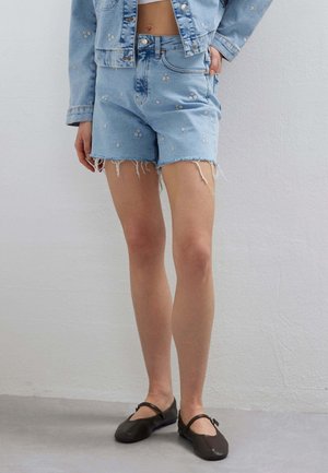 Persona che indossa shorts in denim azzurro chiaro e giacca abbinata con piccoli ricami floreali bianchi, abbinati a ballerine nere Mary Jane.