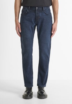 Jean en denim foncé avec une coupe droite. Comprend cinq poches, un zip à boutons et des détails de couture subtils. Portés avec des bottes noires.