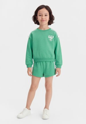 Enfant debout, portant un sweat-shirt vert et un short assorti avec des baskets blanches, souriant devant un fond blanc uni.