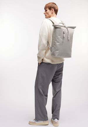 Grauer Rolltop-Rucksack mit verstellbaren Trägern, Reißverschlusdetail und minimalistischen Design. Getragen über einem leichten Pullover und grauen Hosen.