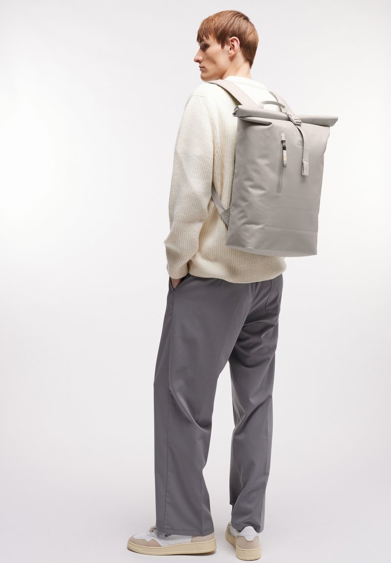 Sac à dos gris avec rabat, bandoulières ajustables, détail de fermeture éclair et design minimaliste. Porté sur un pull léger et un pantalon gris.