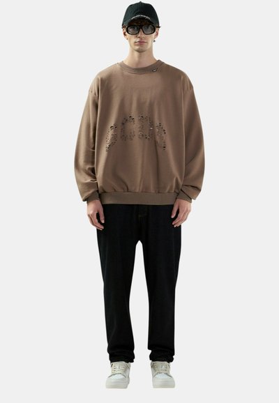 Giovane uomo che indossa una felpa marrone oversize con design traforato, pantaloni neri, sneakers bianche, cappello nero e grandi occhiali neri, in posizione frontale.