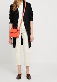 Blazer noir long, chemise blanche, jeans cropped blanc cassé, mocassins marron et petit sac bandoulière rouge avec une poignée sur le dessus.