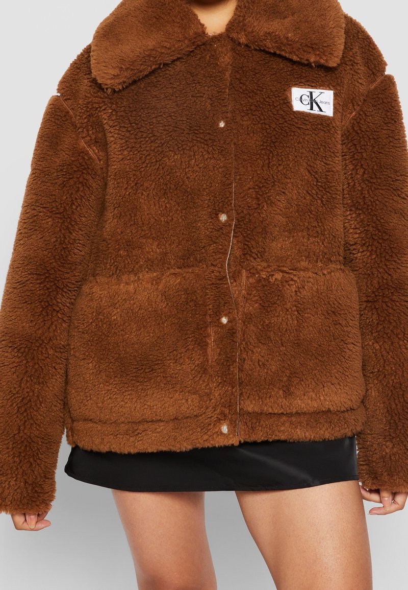 Bruine jas van nepbont met een kraag, knoopsluiting en twee steekzakken aan de voorkant. Heeft een logopatch aan de linkerkant. Zacht, gestructureerd materiaal.