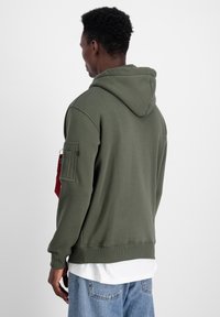 Felpa con cappuccio verde oliva dal taglio rilassato. Presenta una tasca a marsupio, polsini a costine e una tasca laterale per utilità con un accento rosso.