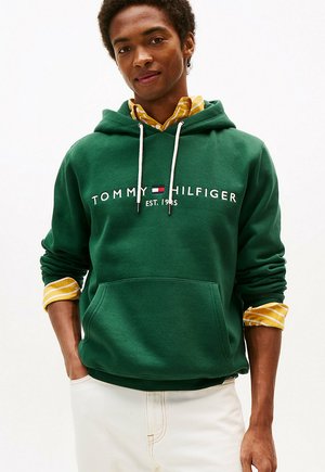 Giovane uomo con una felpa verde Tommy Hilfiger indossata sopra una camicia gialla a righe, con le mani in tasca davanti a uno sfondo bianco semplice.