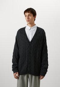 Dark cable-knit cardigan with a V-neck, front buttons, and ribbed cuffs. Worn over a white striped shirt and gray trousers.