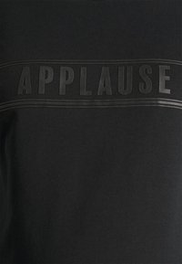 T-shirt de algodão preto com um gráfico em relevo "APPLAUSE" no peito, emoldurado por duas linhas paralelas. Textura de superfície suave.