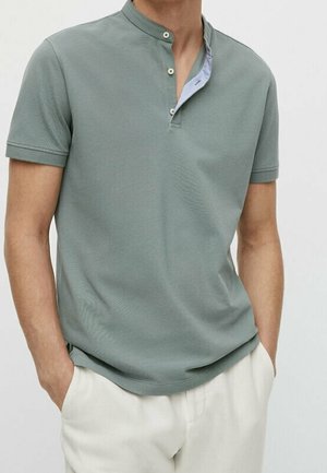 Homme portant un t-shirt henley à manches courtes vert sauge avec trois boutons et un pantalon blanc, mains dans les poches.