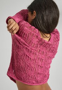 Rosa stickad tröja med vågmönster, lös passform och öppen vävstruktur. Rundad halsringning och något längre bakstycke.