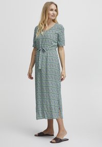 Maxiklänning i grön och blå blommönster, v-ringning, korta puffärmar, knytmidja, sidofickor, lätt tyg, bärs med mörka sandaler.