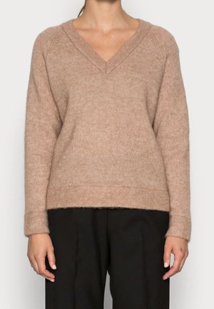 Femme portant un pull duveteux marron clair à col en V avec des manches longues et un pantalon noir ample, montrée du cou jusqu'au milieu de la cuisse.