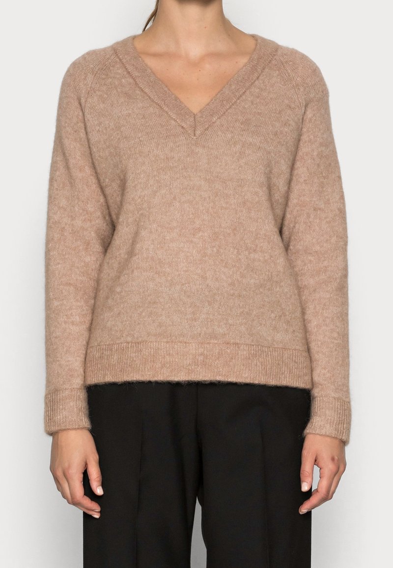 Femme portant un pull duveteux marron clair à col en V avec des manches longues et un pantalon noir ample, montrée du cou jusqu'au milieu de la cuisse.