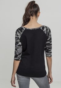 Kvinna med brunt hår i hästsvans, iförd en svart tröja med grå och svart kamouflage, 3/4 ärmar och ljusgrå jeans, står vänd bort.