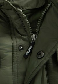 Primo piano di una cerniera di giacca verde oliva con un tirante nero etichettato "OMBRE" e bottoni a pressione metallici visibili.